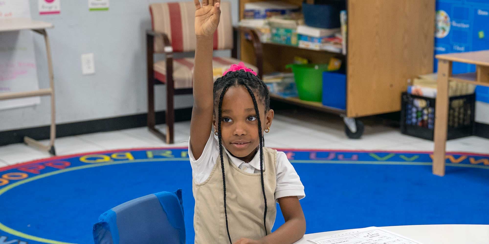 Young student raising a hand at a desk in a classroom.